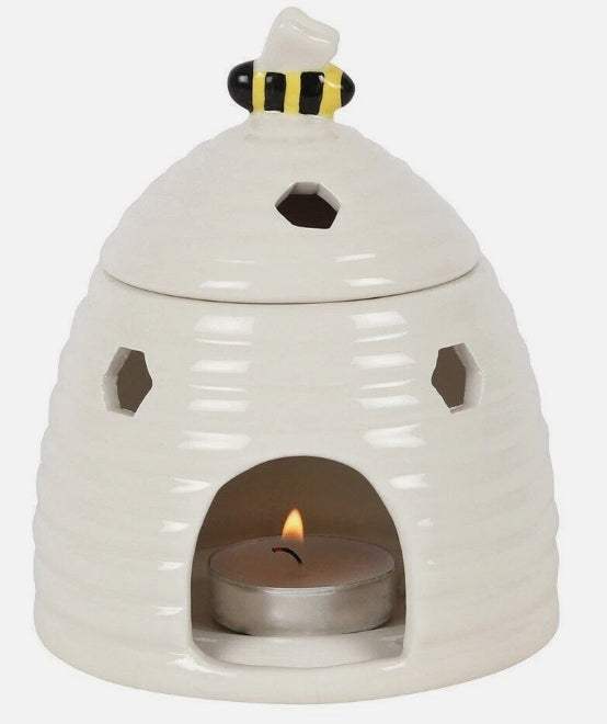 A white ceramic beehive-shaped candle holder with a small tea light candle lit inside, topped with a bee figurine, on a light gray background.