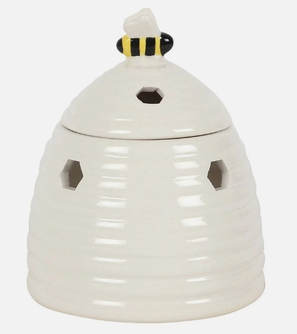 An off-white ceramic beehive-shaped burner with a lid, featuring hexagonal cutouts on the sides and a small bee figure on top of the lid.